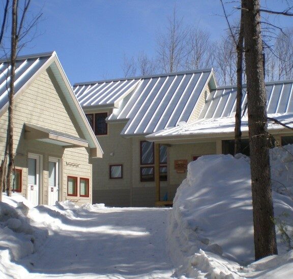 The Poplar Falls Hut with tan wood siding and a metal roof in winter. A cross country ski trail is in the foreground.