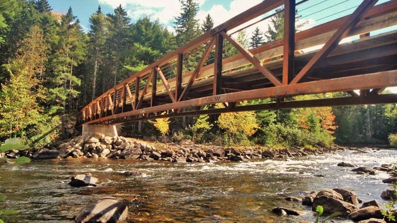 Image of the Chappell Bridge, a steel framed pedestrian bridge with wood decking going over the Dead River near Grand Falls